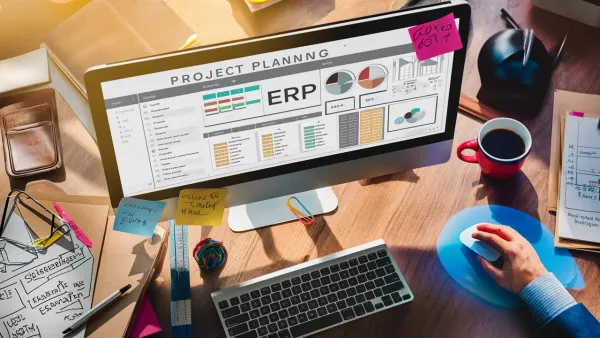 Computador moderno com painel ERP cercado por documentos de planejamento e notas coloridas em mesa de madeira.